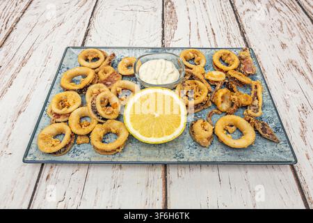 Knusprig gebratene Calamariringe mit Zitronenscheibe und cremiger Dip, arrangiert auf einer blau glasierten rechteckigen Keramikplatte Stockfoto