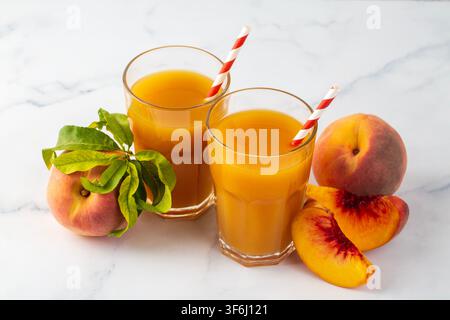 Zwei Gläser frischer Pfirsichsaft mit rot gestreiften Strohhalmen, umgeben von ganzen und geschnittenen reifen Pfirsichen auf weißem Marmorhintergrund Stockfoto