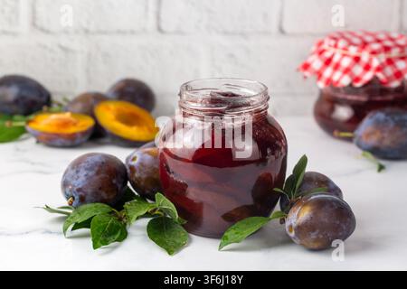 Hausgemachte violette Pflaumenmarmelade in einem offenen Gläser auf weißem Marmorhintergrund, umgeben von frischen reifen Pflaumen Stockfoto
