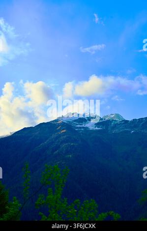 Eine vertikale Landschaftsfotografie eines dunklen, waldbedeckten Berggipfels mit Schneeflecken unter einem lebendigen blauen Himmel, der mit weichen weißen Wolken gefüllt ist. Stockfoto