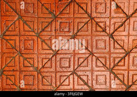 Nahaufnahme von oben auf verwitterte rot-orangefarbene Pflastersteine mit einem erhabenen quadratischen und diagonalen Kreuzmuster, getrennt durch dunkle gerillte Linien Stockfoto