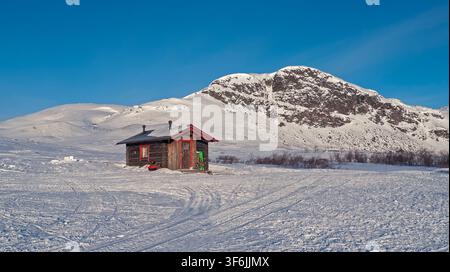 Enontekiö, Finnland - 01.04.2018: Rustikale rote Holzhütte, isoliert in einer schneebedeckten arktischen Landschaft mit felsigen Gipfeln und einem strahlend blauen Himmel Stockfoto