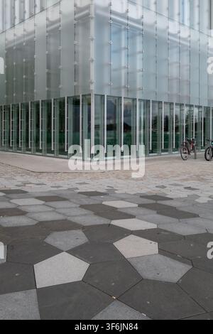 Eckmodernes Glasbürogebäude mit mattierter Fassade, hohen Fenstern und gemusterter Pflasterung in einem leeren städtischen Platz. Saubere architektonische Außenseite mit Stockfoto