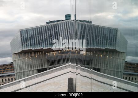 Modernes Glasgebäude, das sich in einer verspiegelten Wand spiegelt, und eine symmetrische städtische Szene unter einem bewölkten Himmel erzeugt. Klare Linien, dramatische Perspektive und zeitgemäß Stockfoto