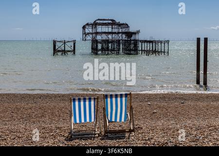 Liegestühle am Kieselstrand in Brighton, mit den Ruinen des West Pier im Hintergrund Stockfoto