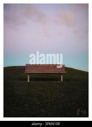 Leere hölzerne Parkbank auf einem grasbewachsenen Hügel unter einem weichen pastellfarbenen bewölkten Himmel im Totara Heights Park, Auckland, Neuseeland Stockfoto