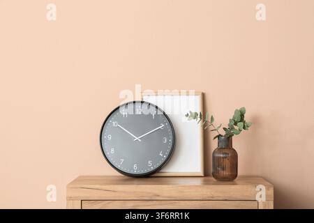 Moderne graue Wanduhr mit einem leeren Bilderrahmen und einem Eukalyptuszweig in einer Vase auf einem Holzschrank, die Gegenstände sind vor einem glatten pfirsichfarbenen Hintergrund platziert Stockfoto