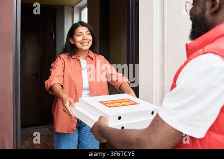 Hispanische Frau, die Pizzakartons von einem schwarzen Kurier an der Tür entgegennimmt Stockfoto
