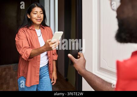 Hispanische Frau, die einen QR-Code auf dem Smartphone eines Lieferanten an der Tür scannt Stockfoto