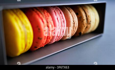 Nahaufnahme bunter französischer Macarons in einer Reihe in einer dunkelgrauen Verpackungsbox, mit solidem blauem Hintergrund. Süße Dessertgebäck-Bäckerei-Lebensmittelphotographie in m Stockfoto