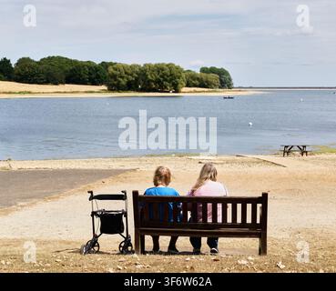 Ein paar Leute sitzen auf einer Bank am Ufer von Rutland Water, England, an einem heißen Sommertag. Stockfoto