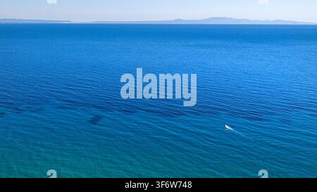 Klares blaues Meerwasser erstreckt sich bis zu einem niedrigen Inselhorizont, während flache türkisfarbene Töne und subtile Wellen ein ruhiges, offenes Meereslandskap schaffen. Stockfoto