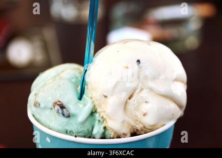 Nahaufnahme von zwei süßen Kugeln cremiger Eiscreme in einem bunten Pappbecher mit einem blauen Löffel. Stockfoto
