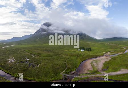 Luftbild von Buchaille Etive Mor, einem Berg in Glen Coe, der teilweise von Wolken verhüllt ist, üppige grüne Hügel, ein gewundener Fluss und ein kleines weißes Haus in Stockfoto