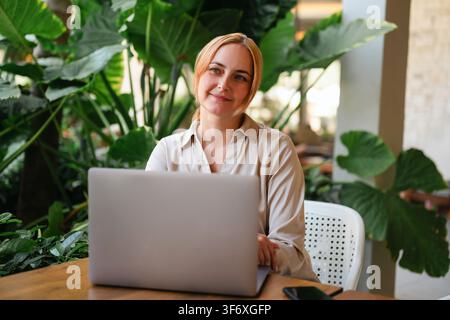Frau arbeitet nachdenklich an einem Laptop in einem grünen Café Stockfoto