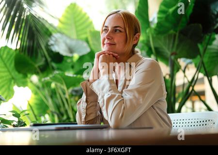 Nachdenkliche Frau am Schreibtisch in einem üppig grünen Café mit Laptop Stockfoto