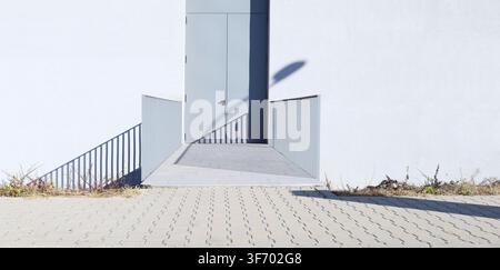 Graue Sicherheitstür an weißer Putzwand mit kleiner metallener Brücke zum Zugang und betoniertem Fliesenpflaster davor. Stockfoto