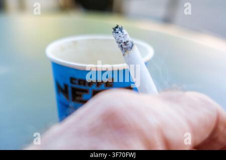 London UK, 07. August 2026, Nahaufnahme der Hand eines Mannes, der eine brennende Zigarette hält, mit einem Caffe Nero-Pappkaffeebecher im Hintergrund Stockfoto