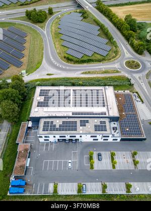 Luftaufnahme eines großen gewerblichen Gebäudedaches, das mit Solarphotovoltaik-Panels bedeckt ist, angrenzend an eine bodengestellte Solarfarm, einen Parkplatz und eine Straße Stockfoto