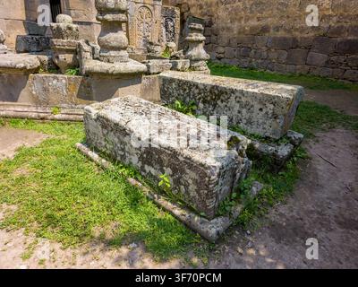 Alte steinerne Grabsteine mit Moos und Gras, das um sie herumwächst. Verwitterte Steinbauten auf einem alten Friedhof. Sonnenlicht scheint auf den moosigen Boden Stockfoto