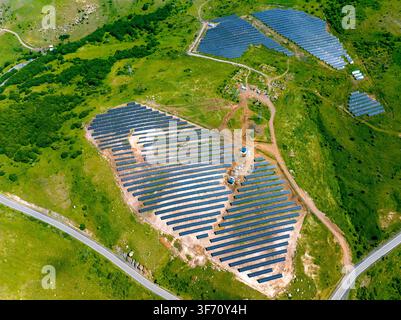 Luftbild von Solarpanelen, die auf grünen Hügeln installiert sind. Die Paneele sind in ordentlichen Reihen über das hügelige Gelände angeordnet. Straßen winden sich um die Anlagen Stockfoto
