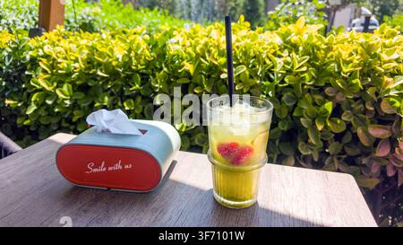 Glas mit gekühltem Getränk mit Strohhalm und Obst. Daneben eine Taschentuchbox auf einem Holztisch. Grünes Gestrüpp im Hintergrund Stockfoto