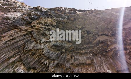 Kanyonwand mit geschichteten Felsformationen unter hellem Sonnenlicht. Vögel fliegen in der Nähe der schroffen Klippenkanten. Natürliche Erosionsmuster sind im Stein sichtbar Stockfoto