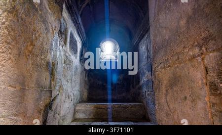 Alte Steintreppe führt nach oben. Sonnenlicht strahlt durch kleines gewölbtes Fenster. Raue, verwitterte Wände umgeben den schmalen Durchgang Stockfoto