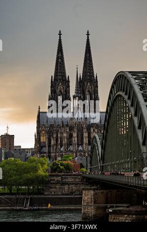 Die gotischen Türme des Kölner Doms erheben sich hinter der Hohenzollern-Eisenbahnbrücke, während Fußgänger bei Einbruch der Dunkelheit über den Rhein gehen. Stockfoto