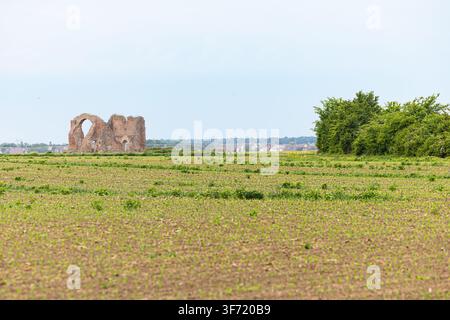 Alte Ziegelruinen stehen allein inmitten eines weiten grünen Feldes mit Hecke und entferntem Dorf am Horizont und rufen Einsamkeit, Geschichte, offene Weite und pa hervor Stockfoto
