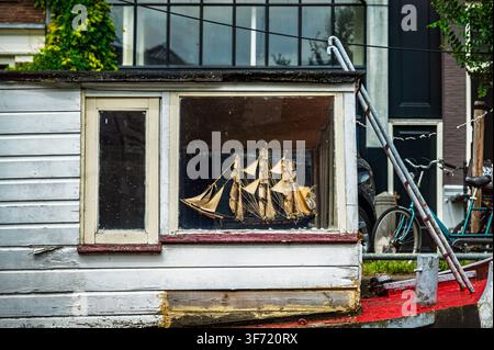 Ein detailliertes Modellsegelschiff steht in der hölzernen Fensterbank eines verwitterten Amsterdamer Hausboots. Stockfoto