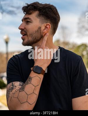 Ein junger bärtiger Mann in einem schwarzen T-Shirt wird im Profil im Freien fotografiert, berührt sein Ohr und schaut zur Seite. Stockfoto