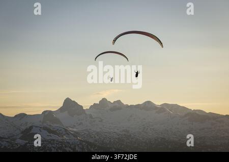 Luftbild von zwei Gleitschirmfliegern, die während der Goldenen Stunde über die schneebedeckten Gipfel des Dachsteinmassivs in Grobming, Steiermark, Österreich schweben. Stockfoto