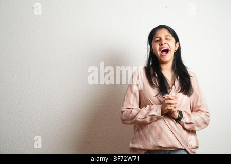 Junge Frau lacht freudig, die Hände zusammengeklammert. Sie trägt eine zarte rosa Bluse und dunkle Hosen. Ihr dunkles Haar fällt locker über ihre Schultern. Rücken Stockfoto