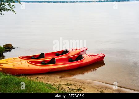 Zwei rote Tandem-Kajaks, die am Ufer eines ruhigen Flusses festgemacht sind. Leere Plastikkanus, die an einem sandigen Strand warten. Friedliche Wassersportausrüstung, bereit für einen S Stockfoto