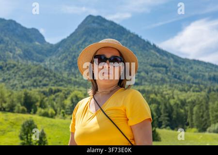 Eine 40-jährige Frau in gelbem Hemd, Sonnenbrille und Strohhut steht vor grünen Alpenbergen Stockfoto