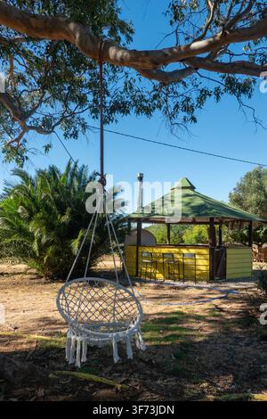 Macrame-Hängestuhl, der von einem großen Ast in einem Hinterhof mit einem gelben Außengarten-Bar-Kiosk, einer Palme und einem klaren blauen Himmel aufgehängt ist. Stockfoto