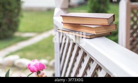 Stapel alter Hardcover-Bücher auf einem Holzzaun in einem Garten mit weichem verschwommenem Grün und blühender pinker Blume, natürliches Licht, friedliches Lesen und Freizeitkonzept im Freien Sommer-Copyspace Stockfoto