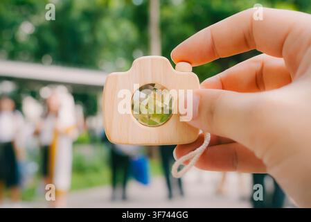 Hand hält hölzerne Spielzeugkamera mit verschwommenem Hintergrund von Absolventen in akademischer Robe, Konzept eines Abschlussfotografie-Dienstes, kreatives Geschäft. Stockfoto