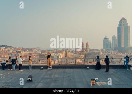 Frau posiert auf einem Hocker an einem Dachaussichtspunkt mit Blick auf die Skyline von Qingdao Stockfoto