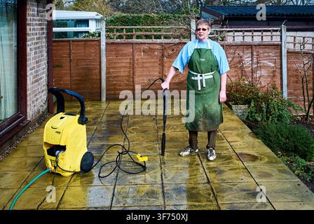 Eine ältere Frau in einer schlammgrünen Schürze hält einen Hochdruckreiniger-Sprüher und reinigt eine nasse Gartenterrasse in einem Vorstadt-Hinterhof in Großbritannien Stockfoto
