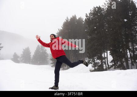 Vollständige Ansicht eines energiegeladenen jungen Mannes in einer roten Jacke, der Spaß hat, als er im Schnee eines nebligen Kiefernwaldes springt Stockfoto