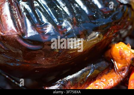 Extreme Nahaufnahme, die sich auf die glänzende und verkohlte Haut einer gegrillten Aubergine mit deutlichen Grillspuren vom Kochen an einer offenen Flamme konzentriert. Stockfoto