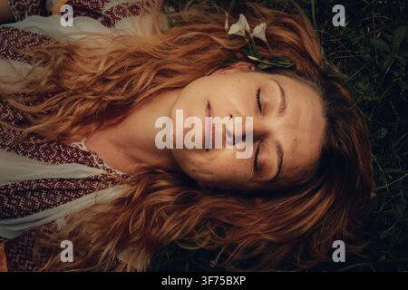 Nahaufnahme-Porträt einer jungen rothaarigen Frau, die eine traditionelle rumänische Bluse trägt und mit geschlossenen Augen im Gras liegt, Kreis Gorj, Rumänien. Stockfoto