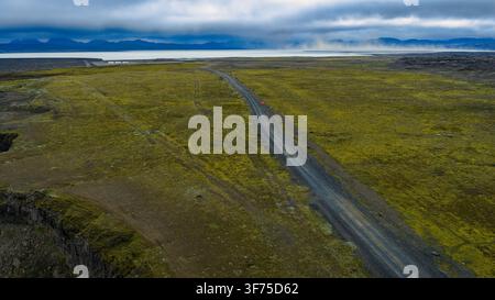 Drohnenperspektive einer asphaltierten Straße, die durch moosbedecktes Basaltermittel in Südisland schneidet, mit Gletscherbergen am Horizont sichtbar. Stockfoto