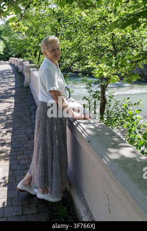 Elegante Seniorin in lässiger Kleidung, die sich an einem steinernen Geländer auf einer Flusspromenade in einem grünen Park an einem sonnigen Sommertag lehnt. Stockfoto