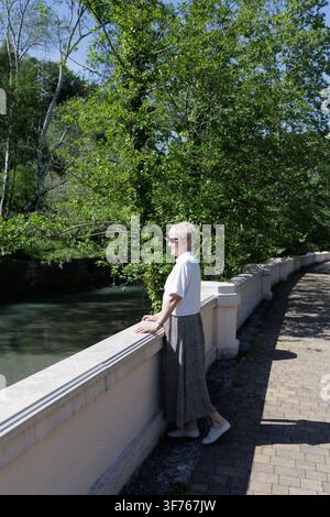 Intensive reife Frau mit kurzem Haar, die eine Sonnenbrille trägt und nahe einer Steinbalustrade auf einer Parkpromenade steht und wegschaut. Stockfoto