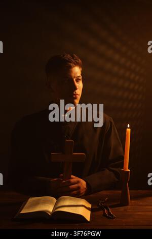 Priester hält Kreuz an hölzernem Tisch mit Heiliger Bibel, Rosenkranz und Kerze vor dunklem Hintergrund Stockfoto