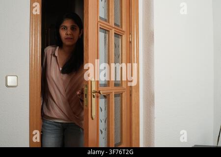Junge Frau steht in der Tür und schaut nach vorne. Ihr Ausdruck ist neutral, ruhig und nachdenklich. Sie trägt ein weiches, strukturiertes Oberteil und Jeans. Holztür Stockfoto