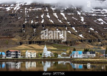 Seyðisfjarðarkirkja blaue Kirche und Stadtgebäude, die sich in ruhigem Wasser unter schneebedeckten Bergen in Seyðisfjörður im Osten Islands spiegeln Stockfoto